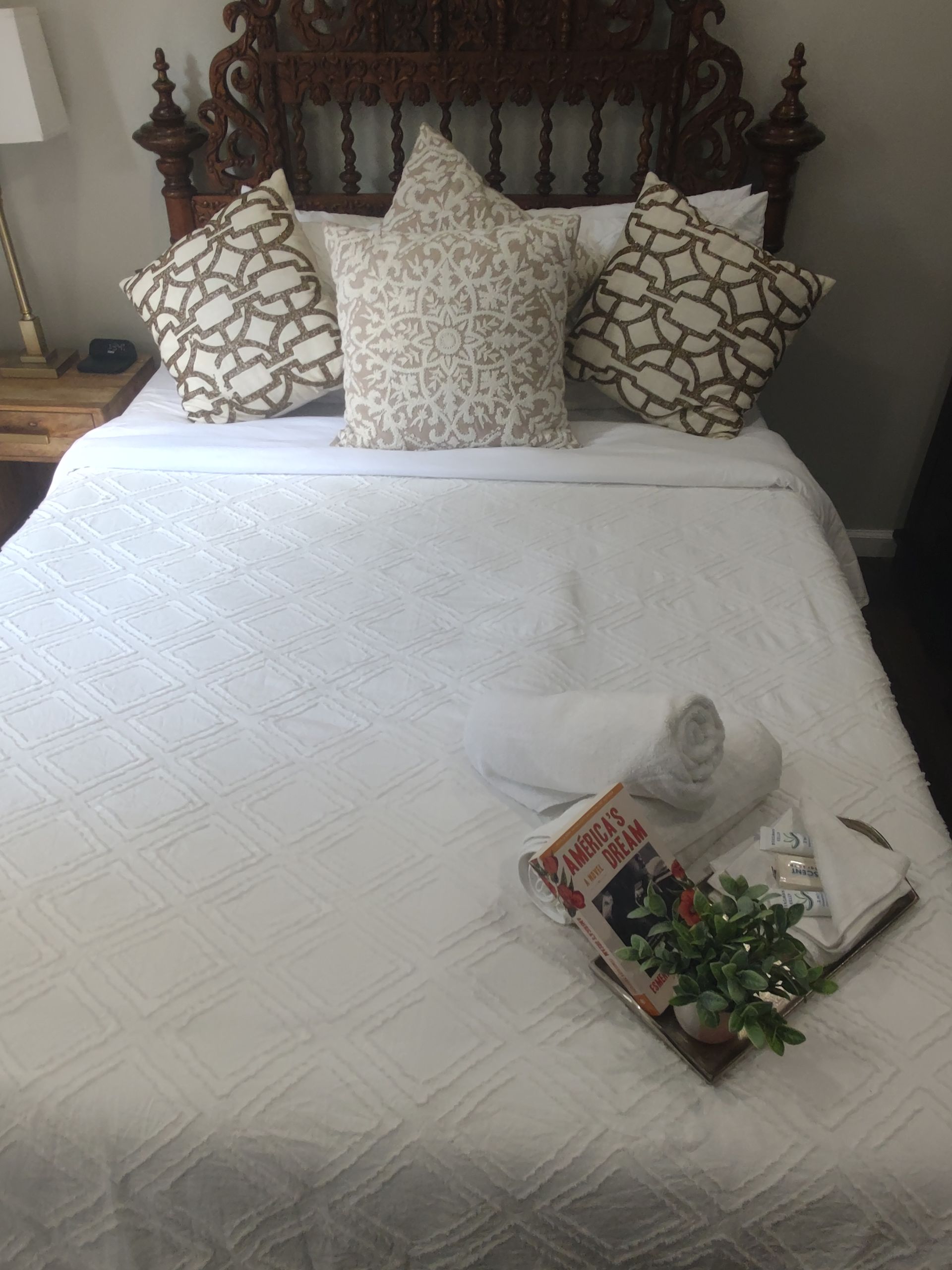 Bedroom with ornate wooden headboard, white bedding, decorative pillows. Book and greenery on bed.