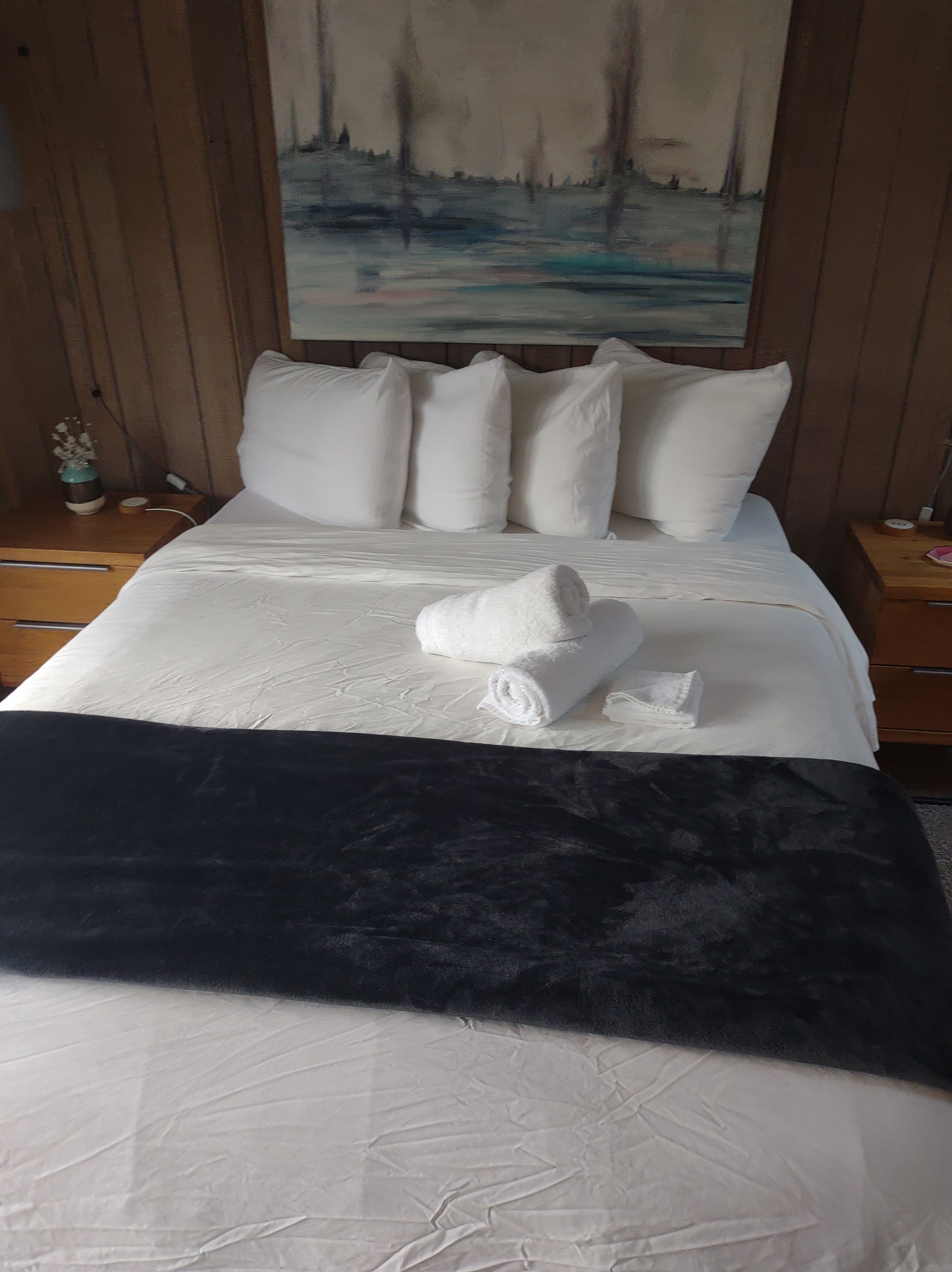 Bedroom with a bed made with white linens, pillows, and rolled white towels. A black throw and artwork hang above.