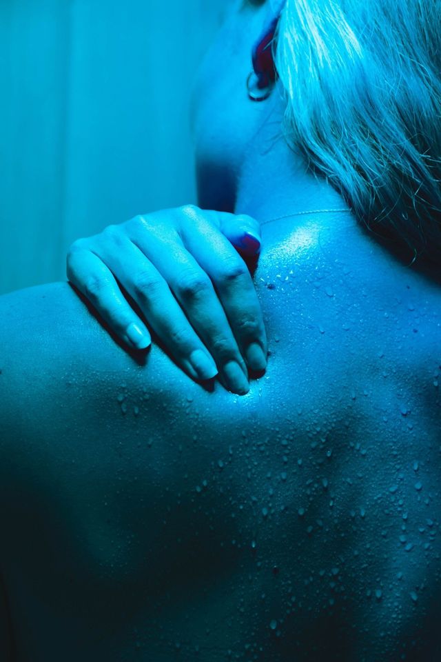 Blue-lit close-up of a person’s bare back with a hand resting on the shoulder