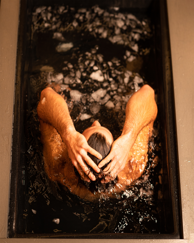 Person lying in a dark tub filled with water, hands over face, with white foam on the surface