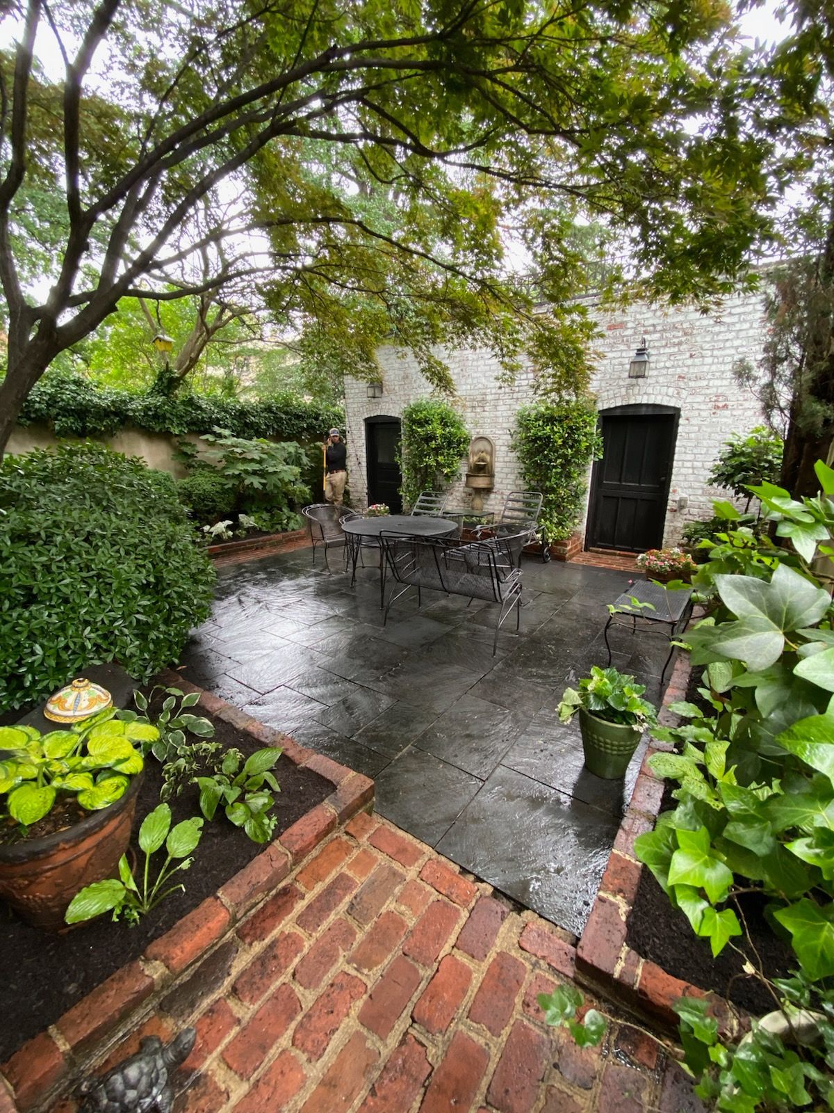 Brick-lined patio with wrought iron table and chairs, surrounded by greenery and brick walls.