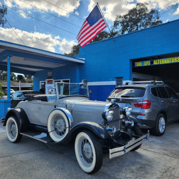 Vintage silver roadster parked outside a blue auto shop with an American flag overhead