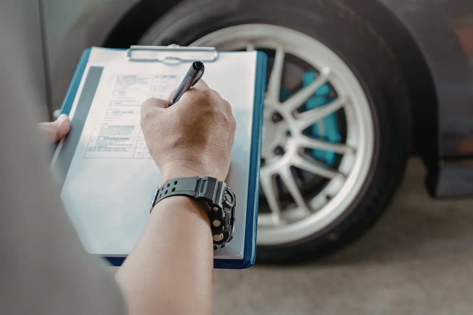 Person writing on a clipboard beside a car wheel and tire inspection.