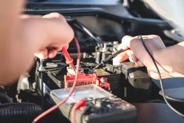 Hands using red and black jumper wires to test a car battery and electrical components under the hood