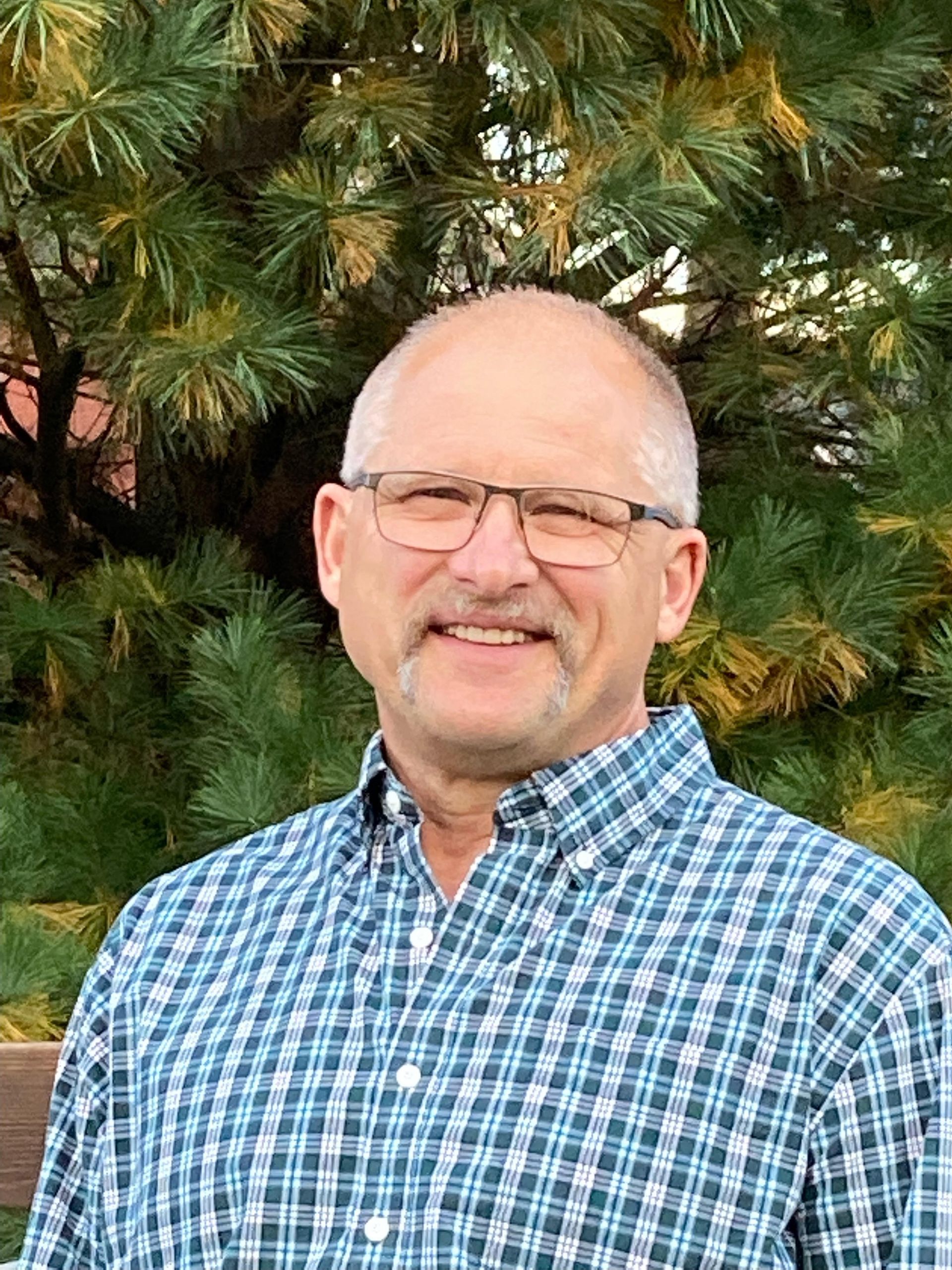 A man wearing glasses and a plaid shirt is smiling in front of a pine tree.