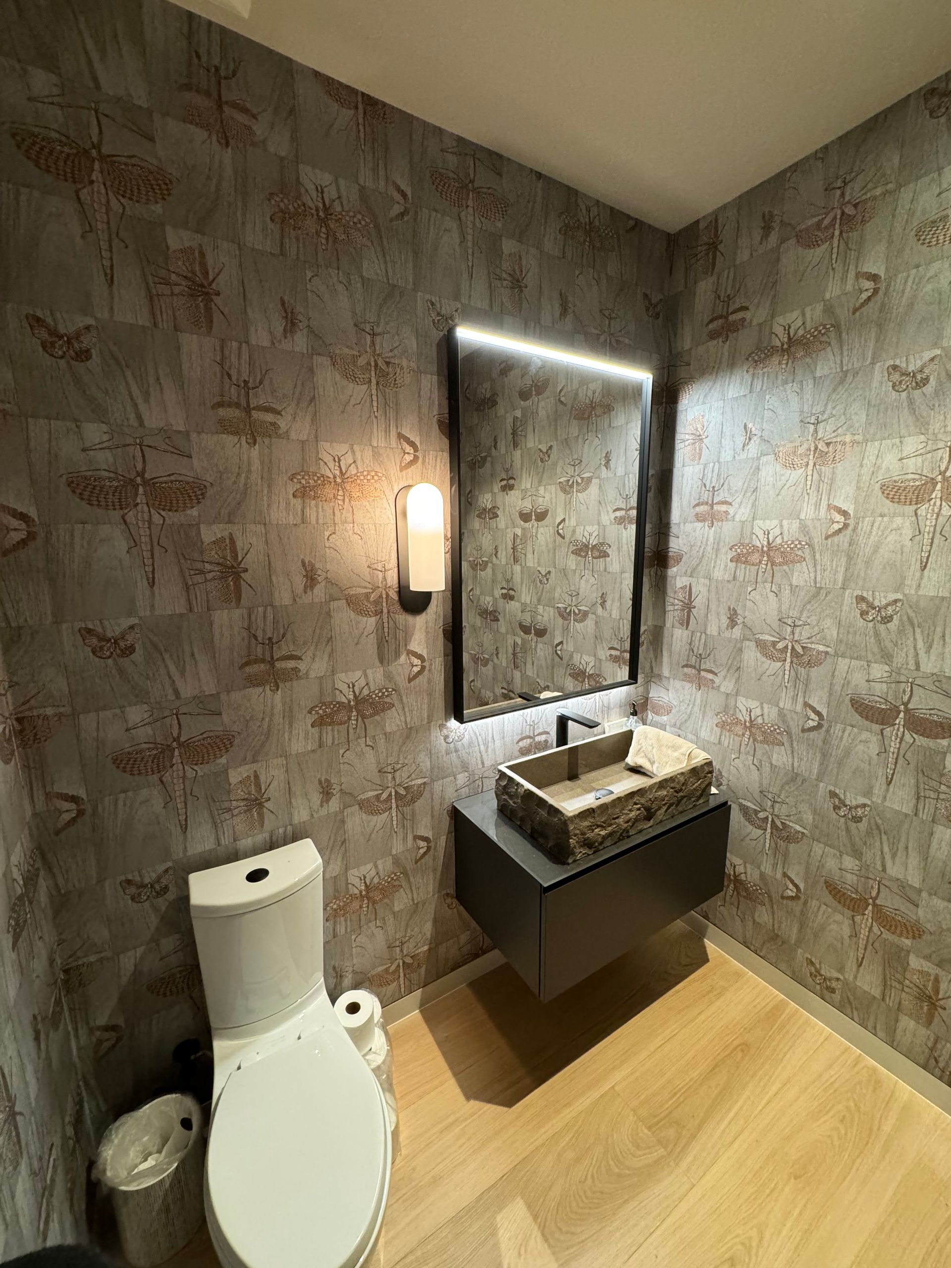 Small bathroom with toilet, wall sink, mirror, patterned wallpaper, and wood floor.
