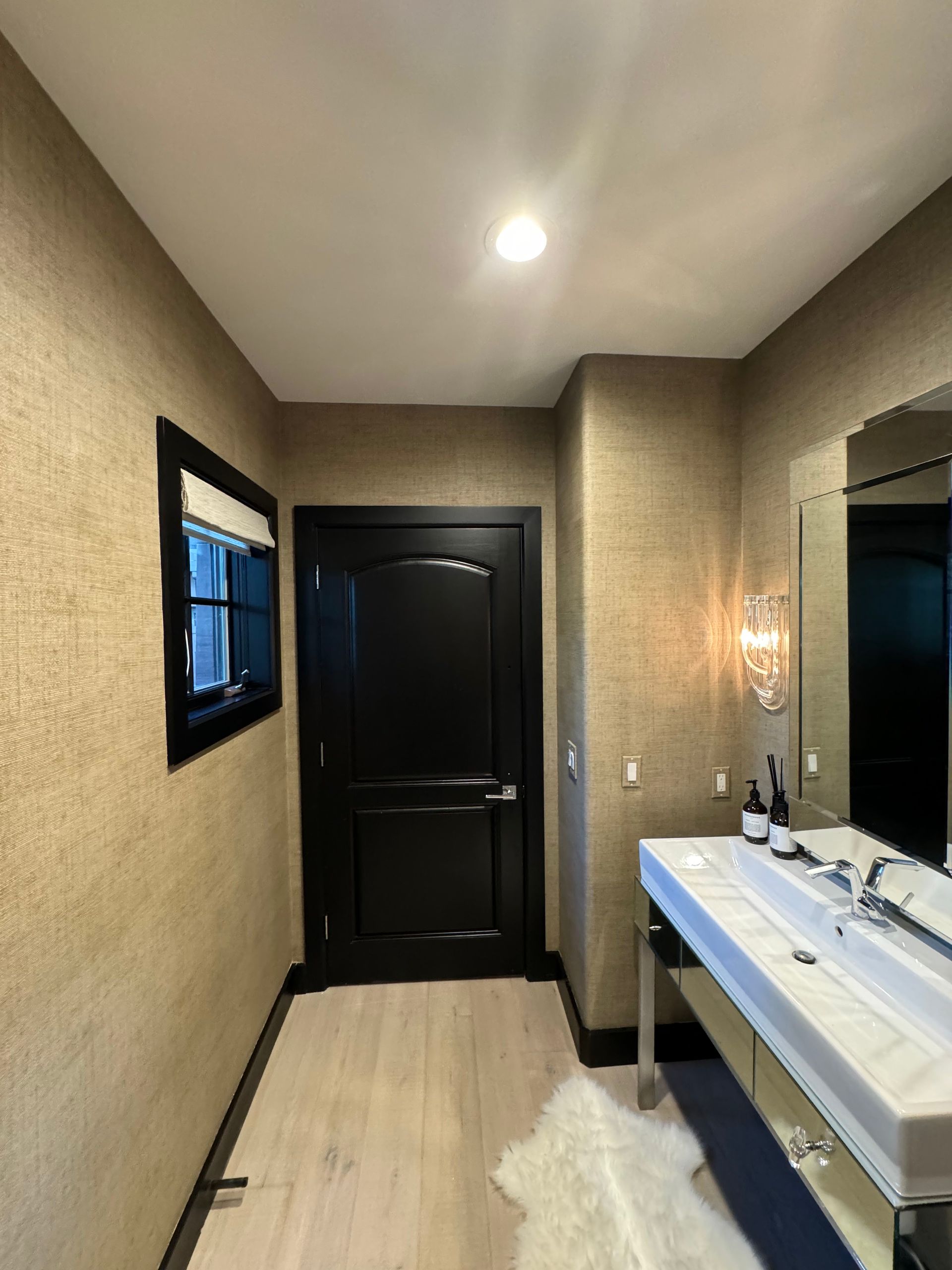 Narrow hallway with beige walls, black door, ceiling light, and a white countertop with sinks on the right.