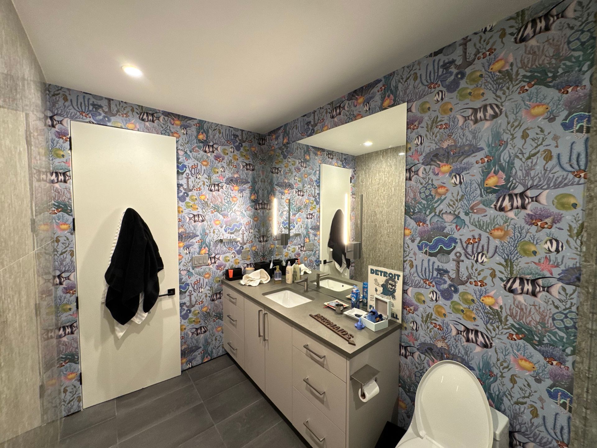 Bathroom with fish-themed wallpaper, double vanity, mirror, and white toilet in a small room