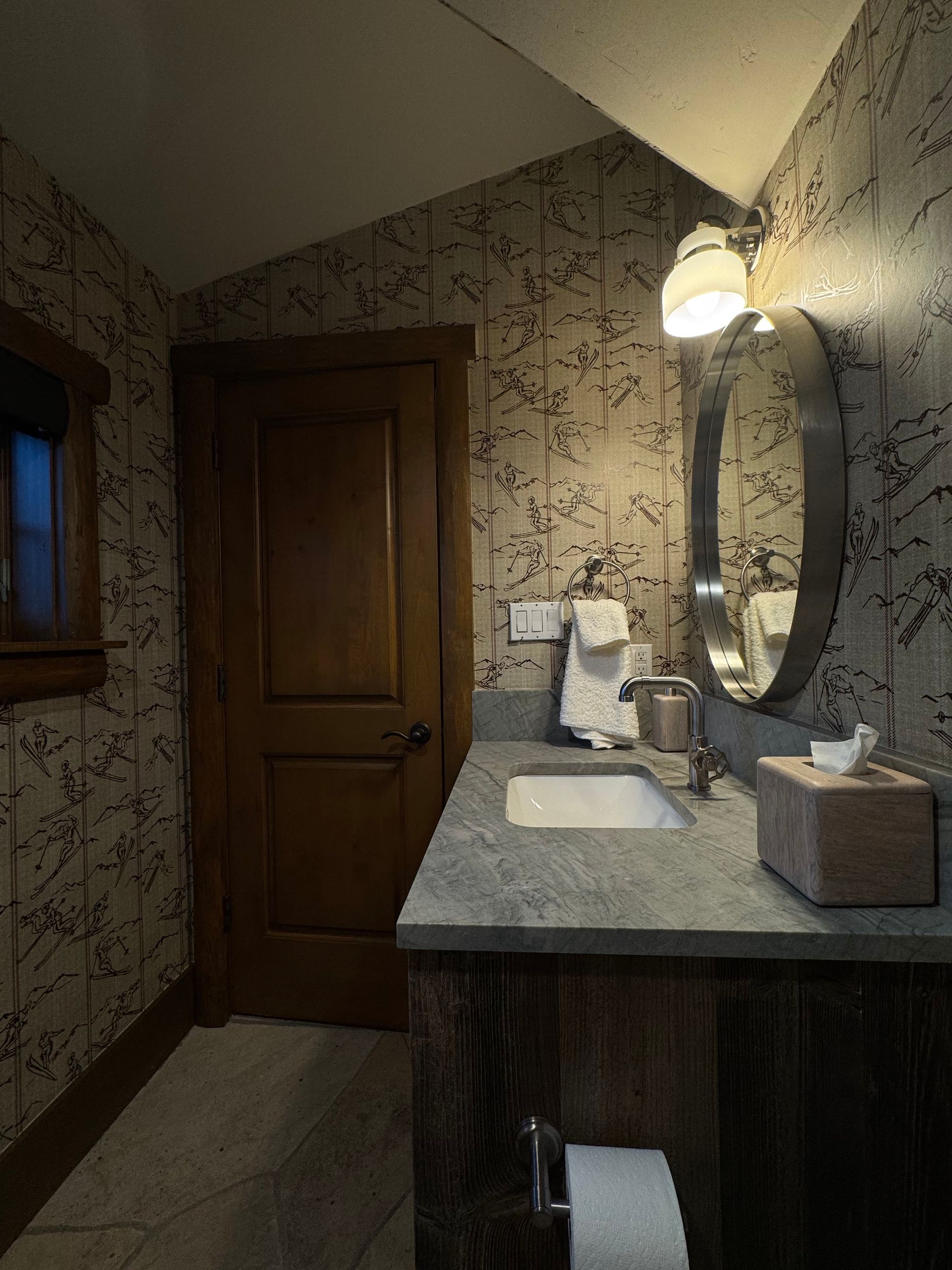 Small bathroom with patterned wallpaper, wood door, vanity sink, mirror, and wall light.