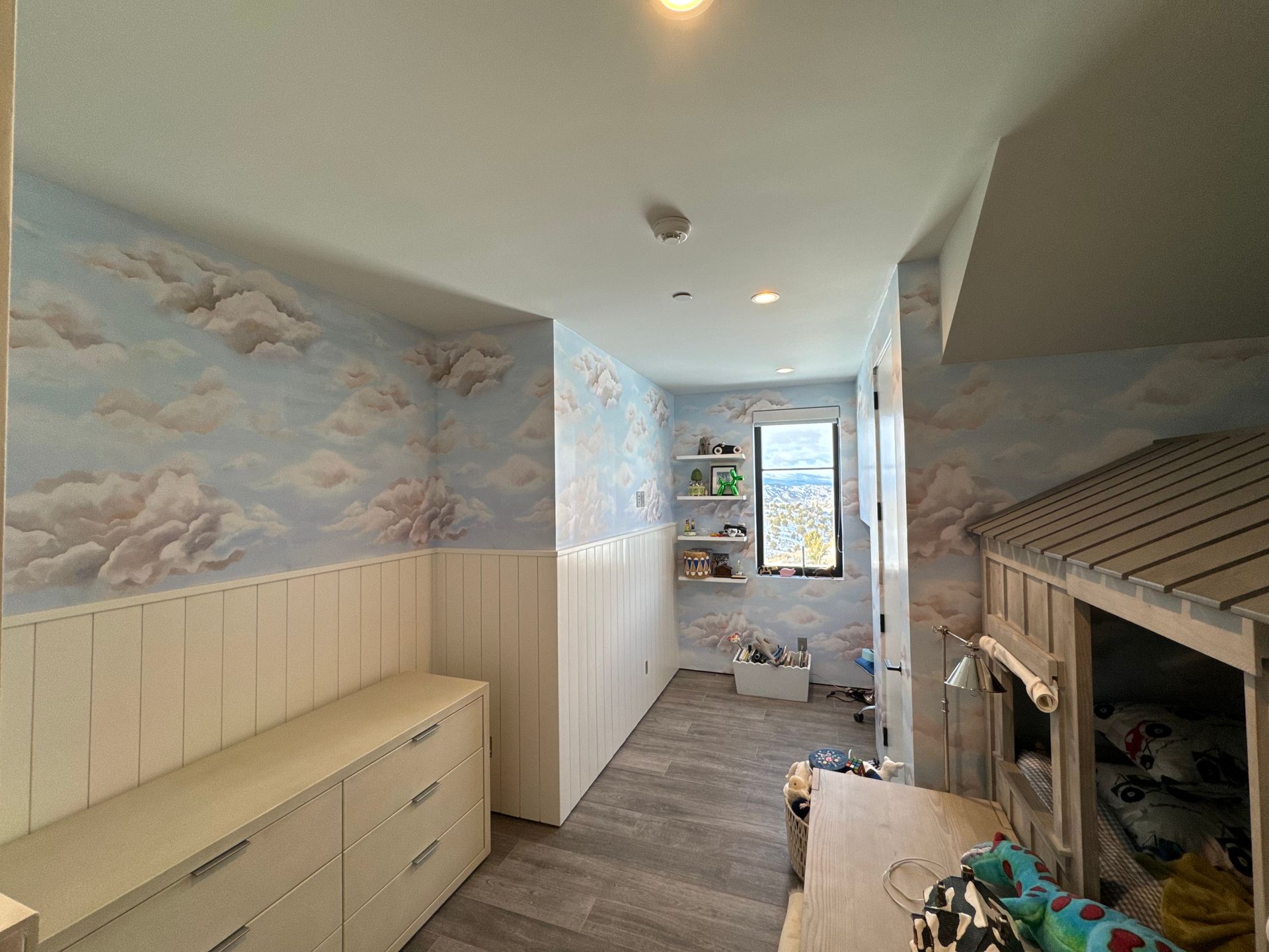 Narrow hallway with cloud mural walls, white wainscoting, dresser, and a bunk bed on the right