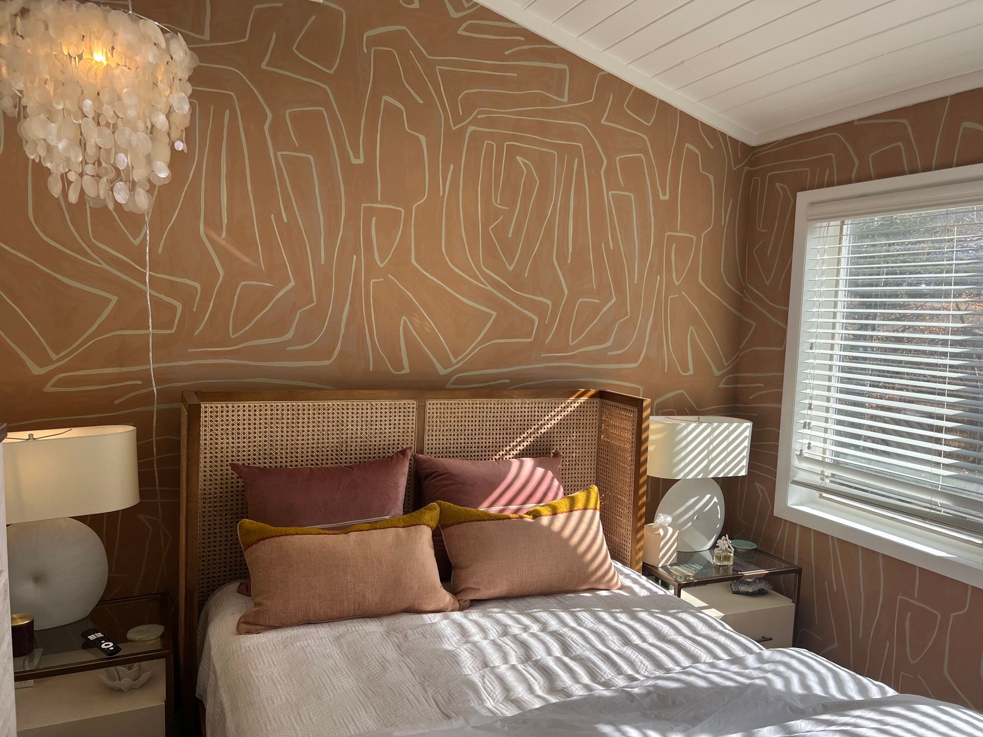 Cozy bedroom with tan patterned walls, woven headboard, white bed, and lamps by a window with blinds