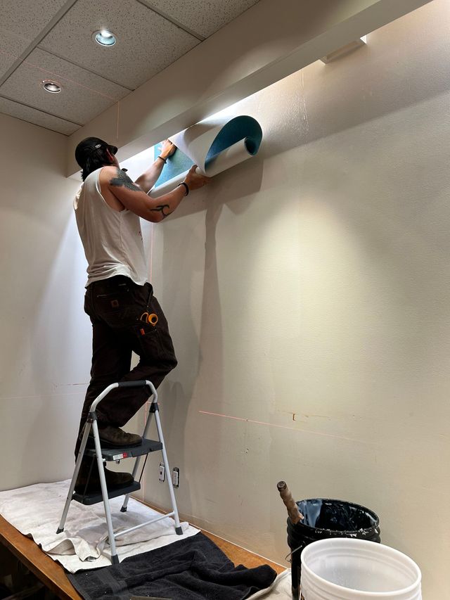 Person on a step ladder painting a wall near the ceiling in a room with drop ceiling tiles.