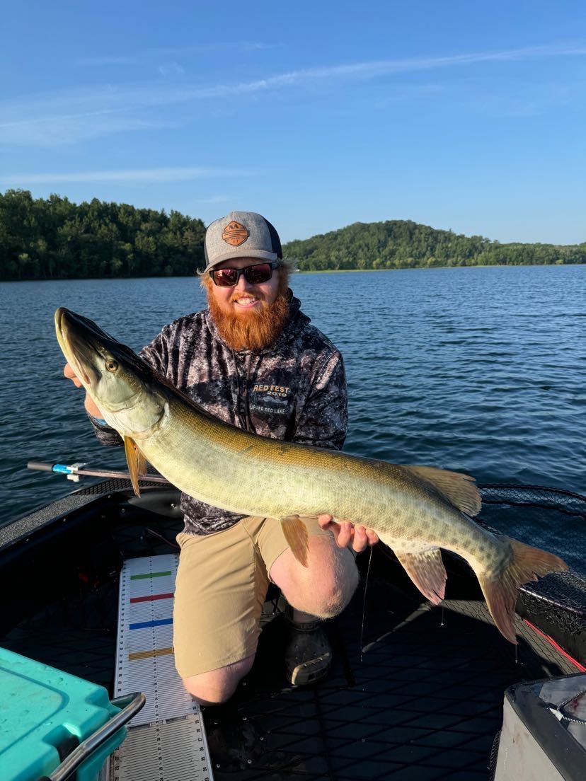 Man holding a large fish on a boat, lake in background, sunny day.