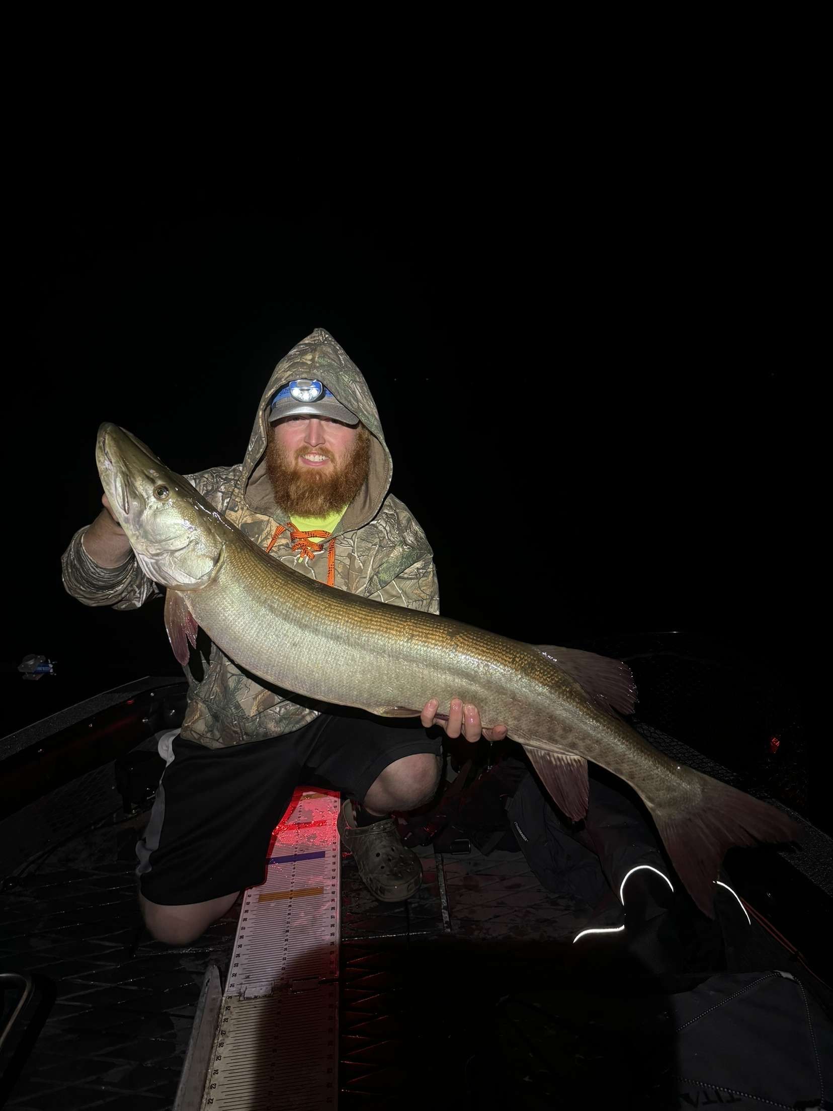 Man in camo holds large muskie fish on a boat at night.