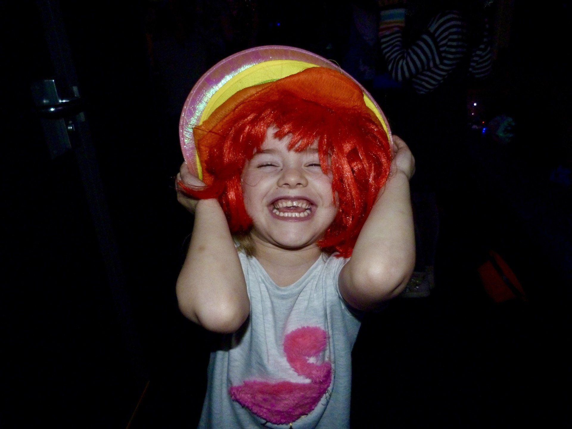 A little girl wearing a red wig and a rainbow hat.