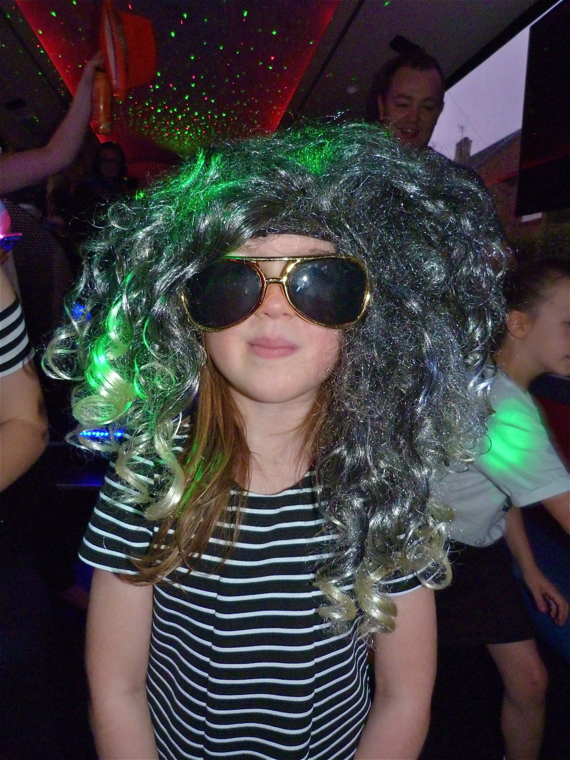 A little girl wearing a wig and sunglasses is standing in a dark room.