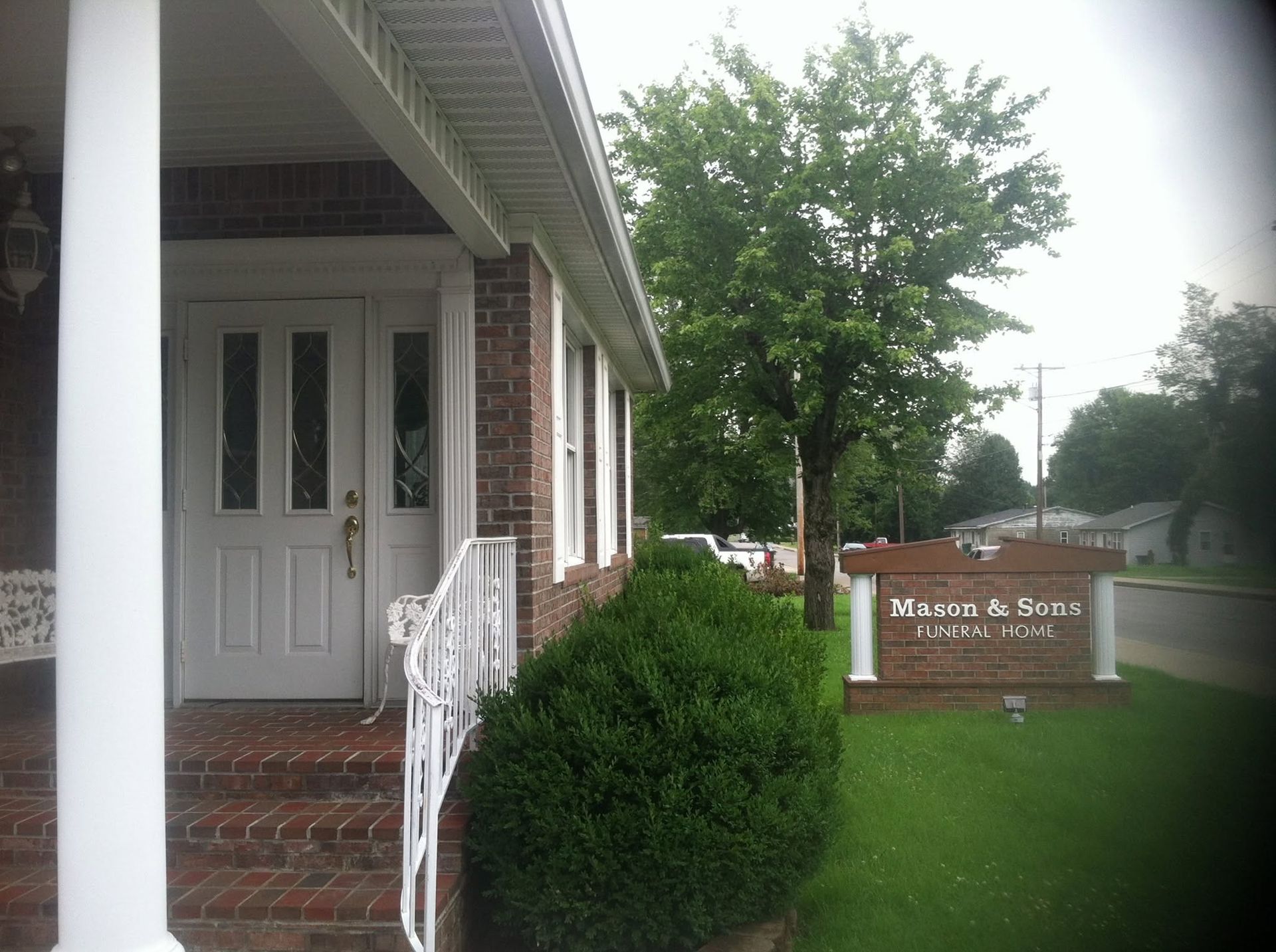 Entrance to a brick building with white doors, shrubs, and a nearby sign by the road.