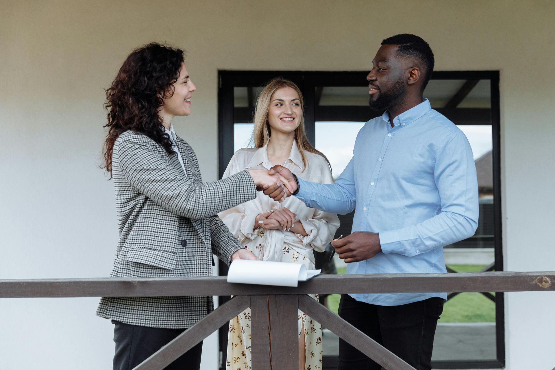 Two people shaking hands, with a woman looking on, outside a home.