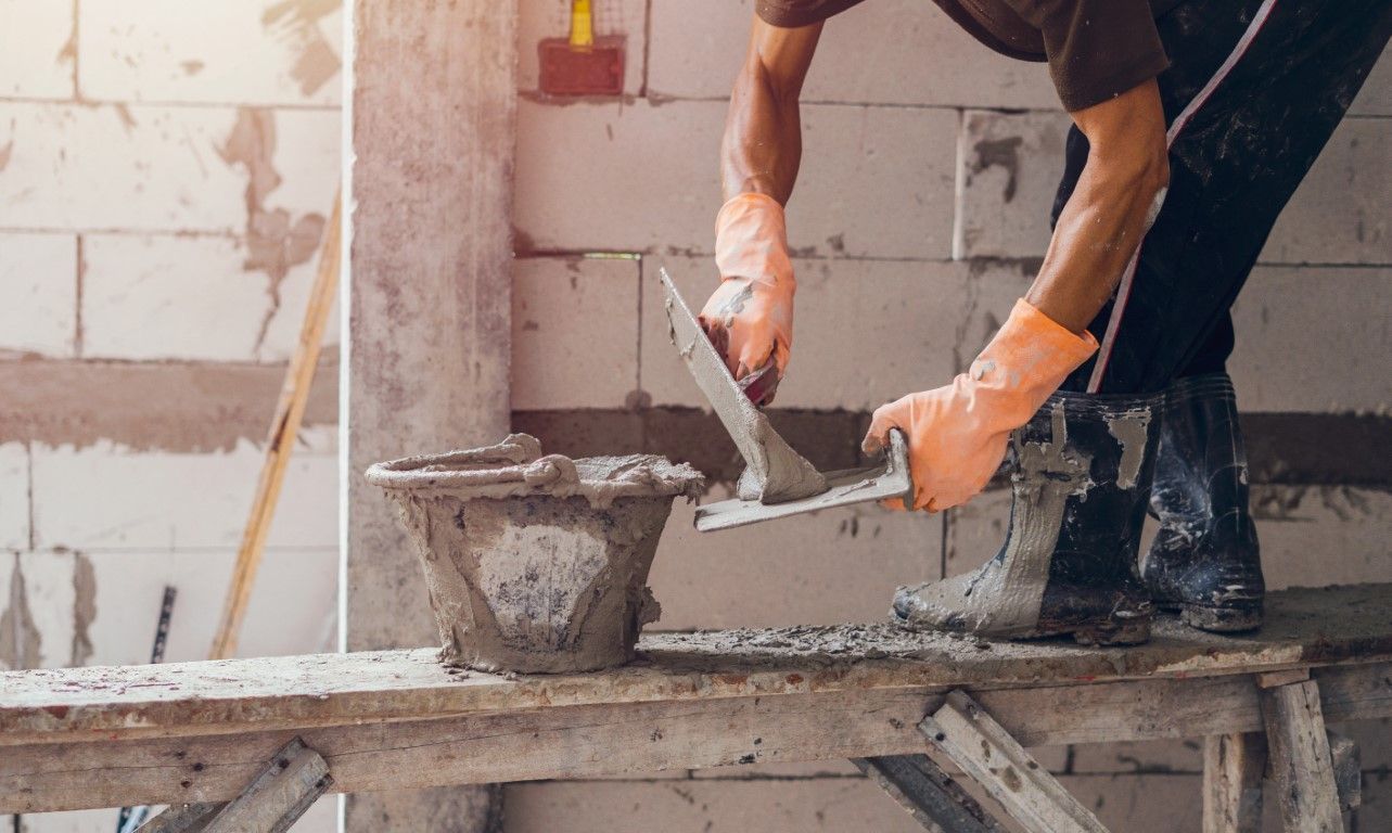 Construction worker spreading mortar on a brick wall with a trowel, wearing orange gloves and boots.