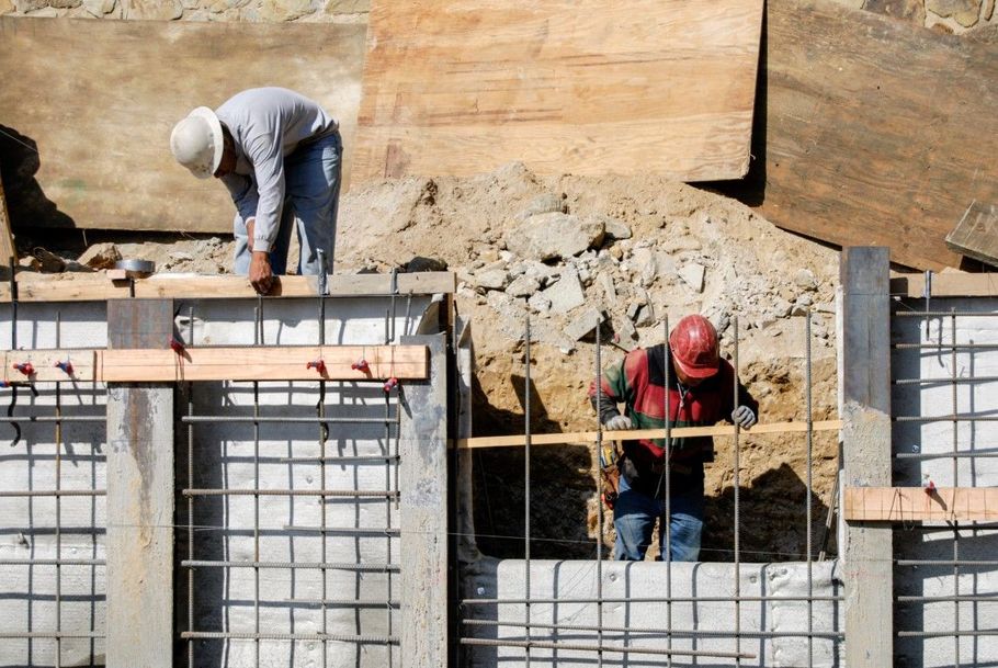 Construction workers reinforcing a concrete wall with rebar and wooden forms at a worksite.