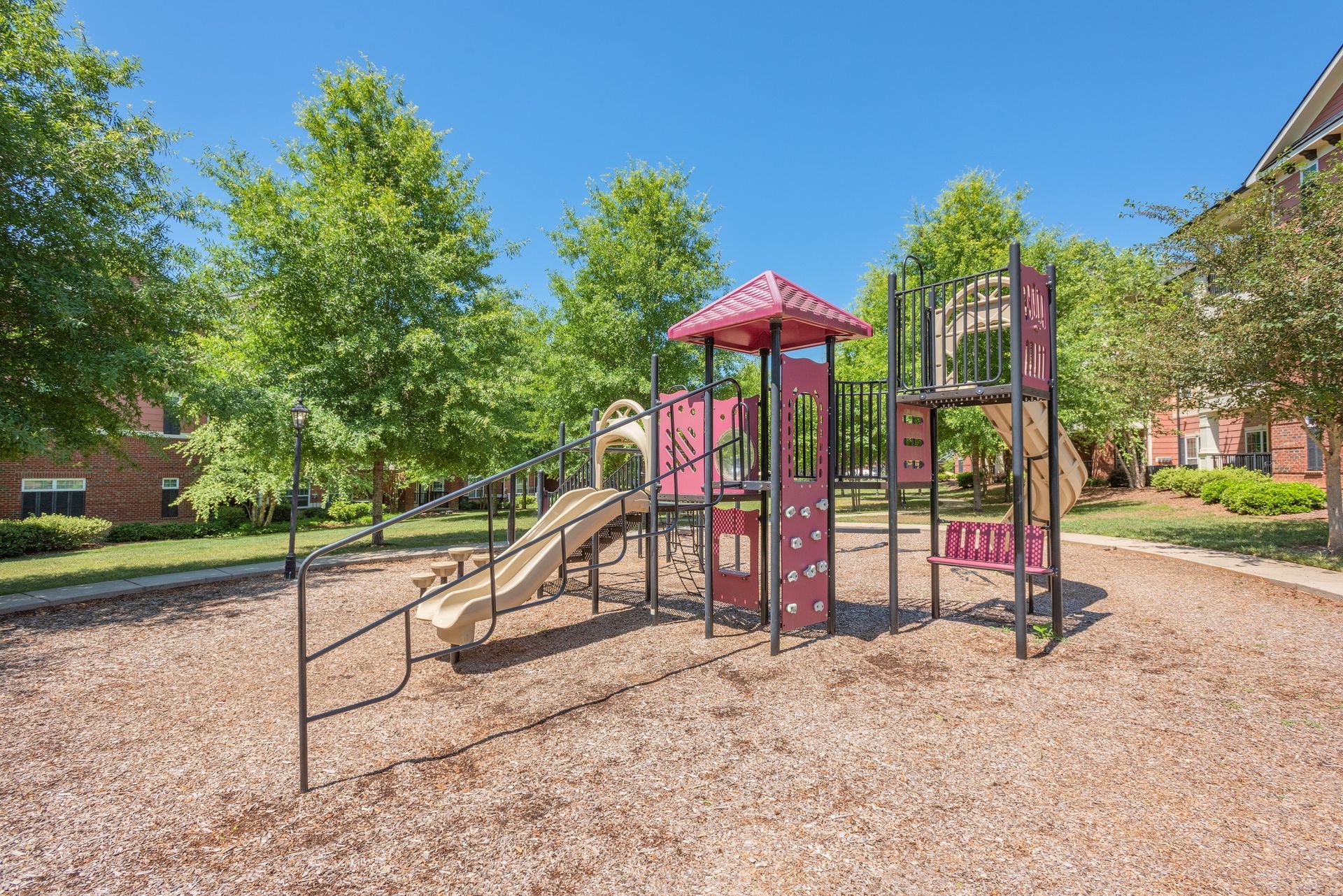 A playground with a multi-level climbing structure, red accents, a slide, and wood chips, set against trees and blue sky at The Vive at Kellswater.