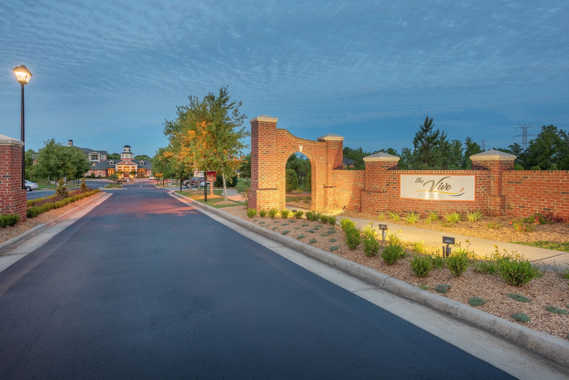 Entrance to a neighborhood with a brick wall, an arched gateway, and a sign, lit by streetlamps at dusk at The Vive at Kellswater.