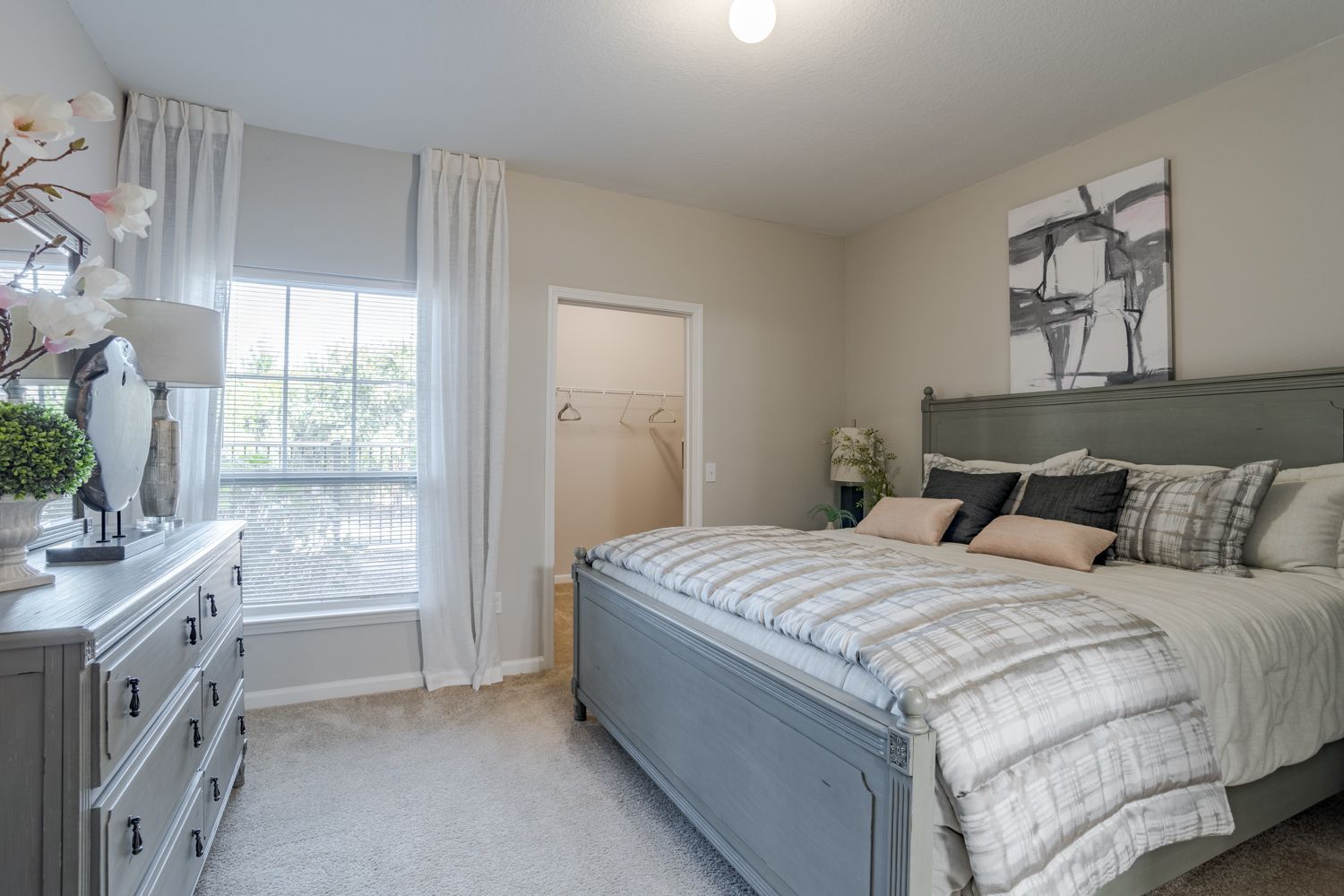 Bedroom with a gray bed, dresser, and wall art. White curtains and neutral walls at The Vive at Kellswater.