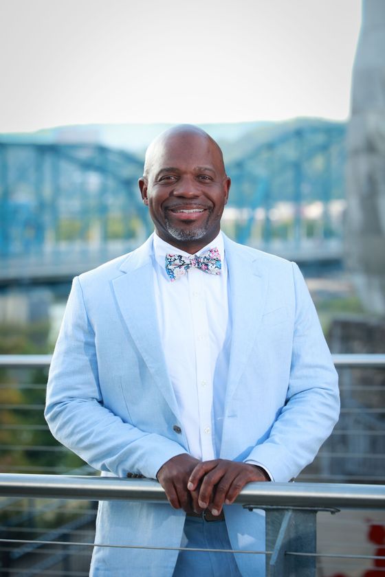 Carlos in a light blue suit and bow tie is leaning on a railing.