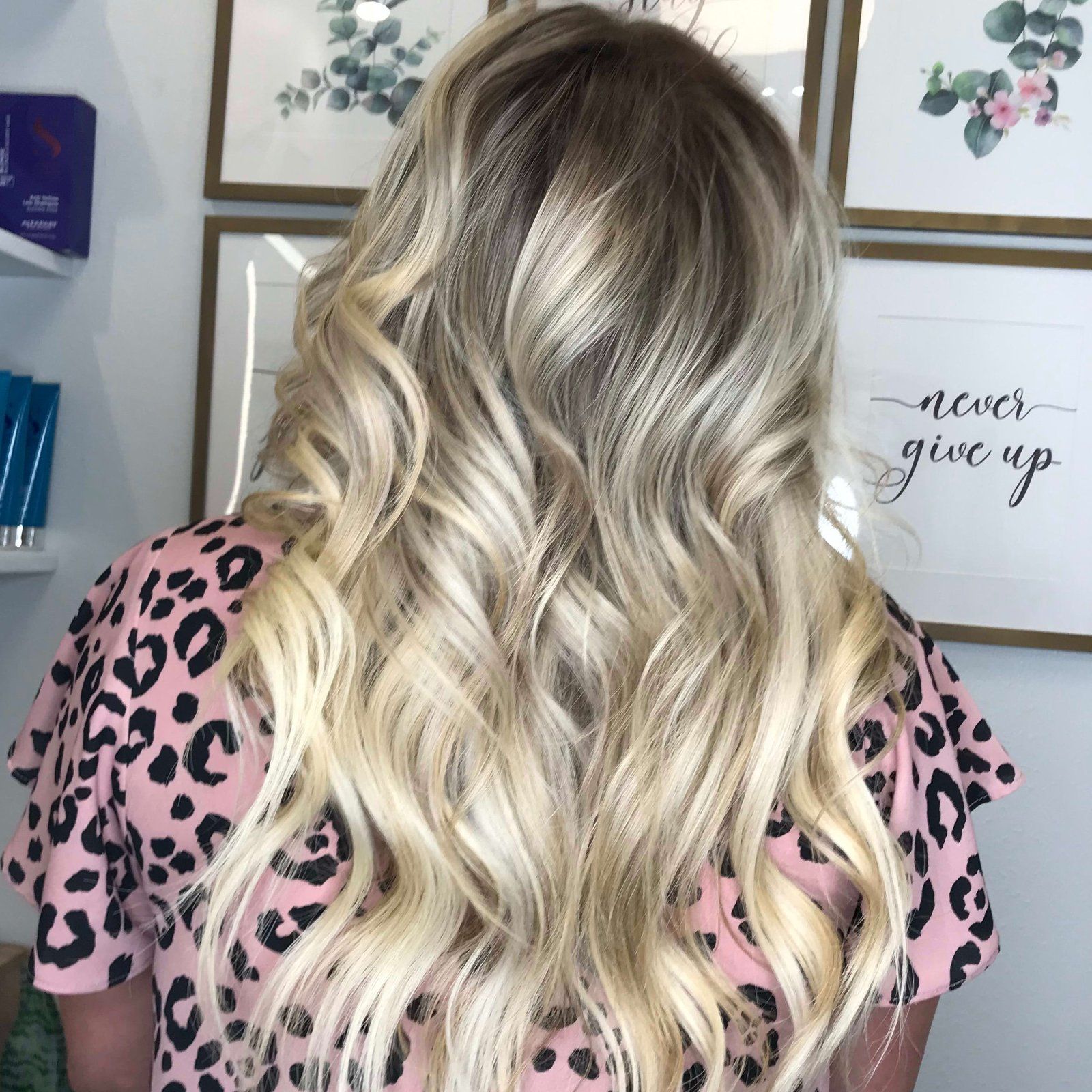 Woman with wavy, blonde-toned hair, wearing a pink leopard-print shirt, in a salon setting.