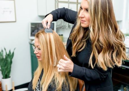 Hair stylist styling a client's hair with a comb. The client has blonde highlights. Brightly lit salon setting.