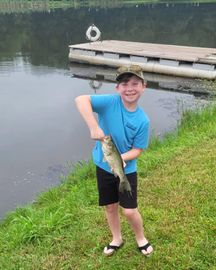 Smiling child holding a fish by a lakeside dock, wearing a blue shirt and black shorts.