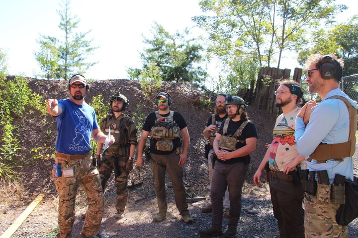 A man in blue gestures to a group of armed hikers standing in a sunny wooded area.