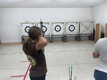 Two people aiming bows at targets in an indoor archery range.