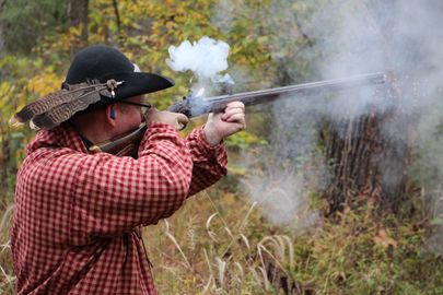 Person in red plaid shirt aiming a smoke-covered rifle outdoors in a wooded area