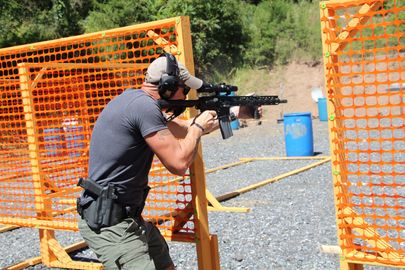 Shooter aiming a handgun past blue barrels beside an orange safety barrier outdoors