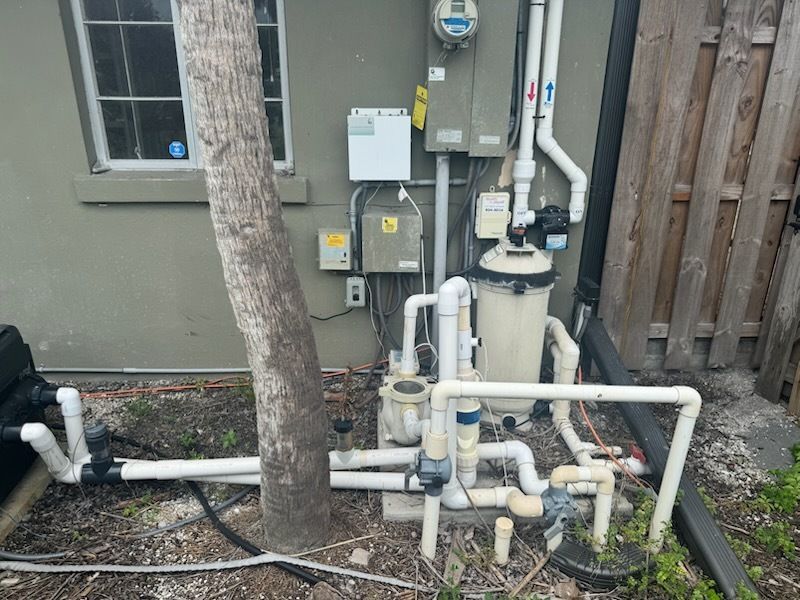 Outdoor pool equipment and plumbing beside a house wall, with a tree in front.