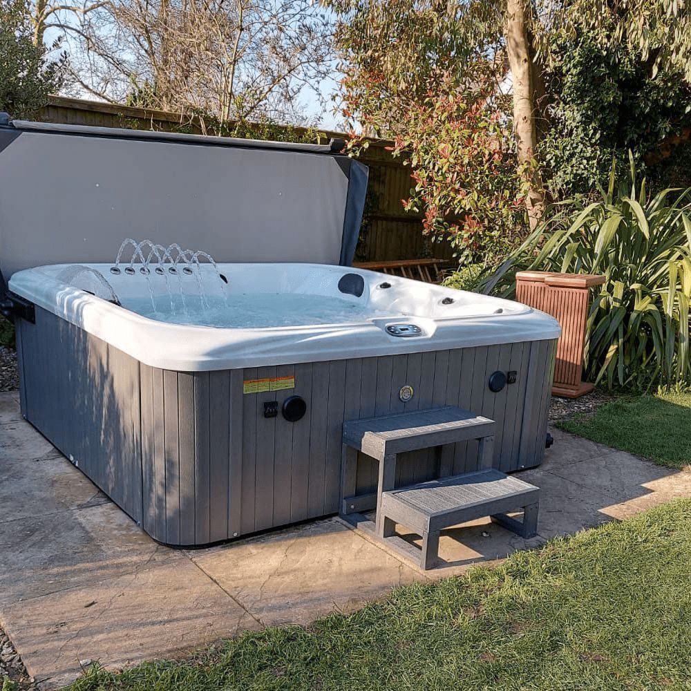 Hot tub outdoors on a concrete pad with two-step stairs. Cover is open. Surrounded by grass, trees, and a wooden fence.
