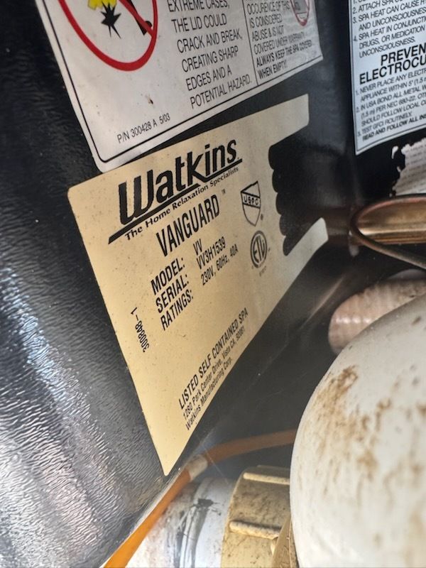 Close-up of Watkins label on a black appliance or surface, with metal knobs and a small pan nearby.