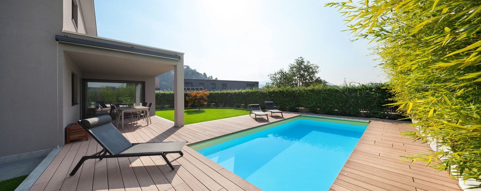 A backyard with a pool, patio, and lounge chair. The sunny setting has lush greenery and a light-colored house.