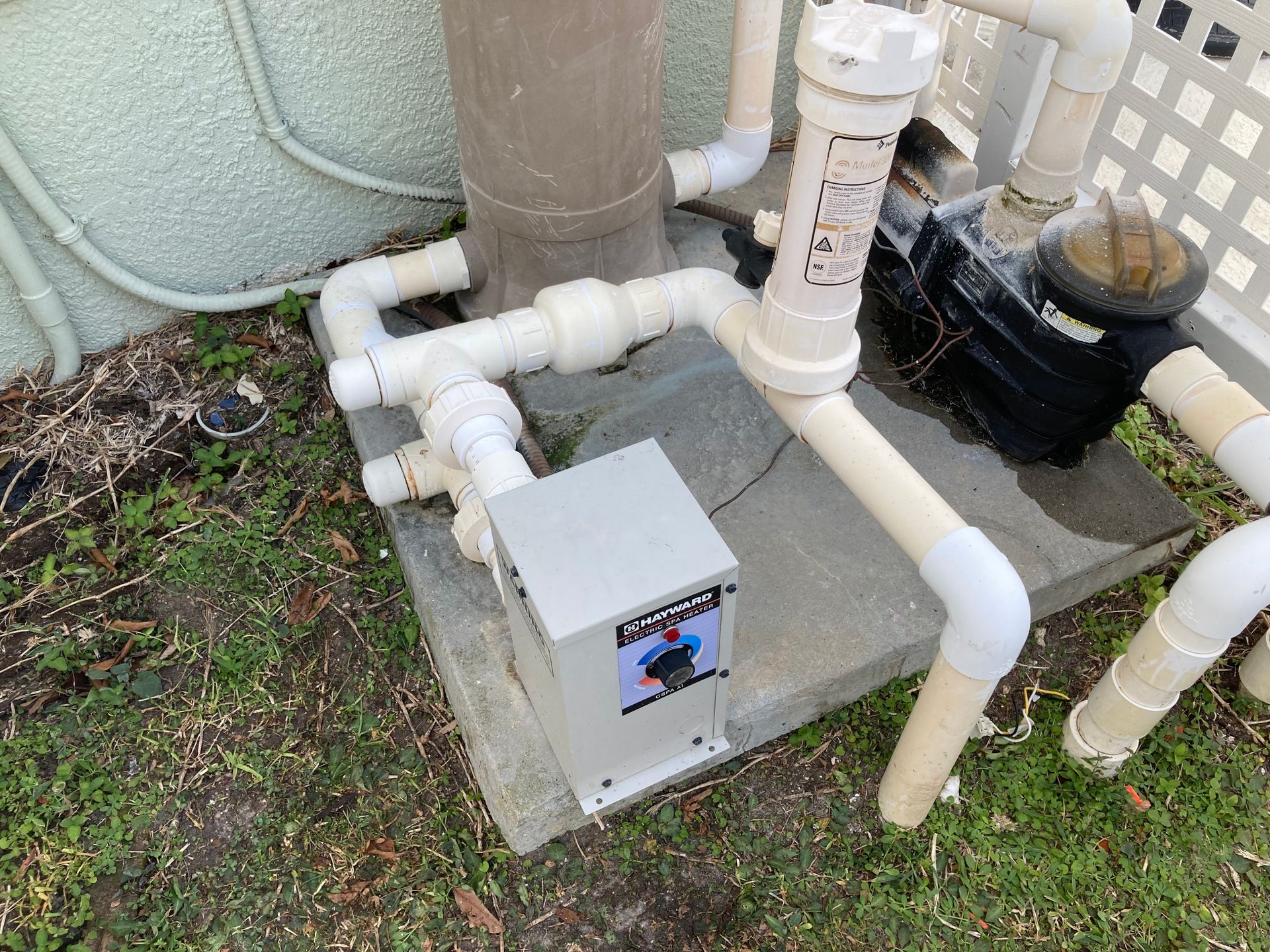 Pool pump system with pipes and a heater on a concrete base next to a wall.