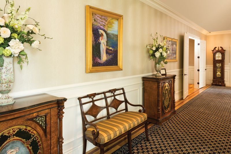 Hallway with floral arrangements, paintings, bench, and grandfather clock.