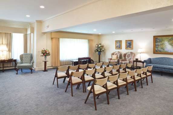 Funeral home interior: chairs arranged for a service, floral arrangements, paintings, windows, and a blue sofa.