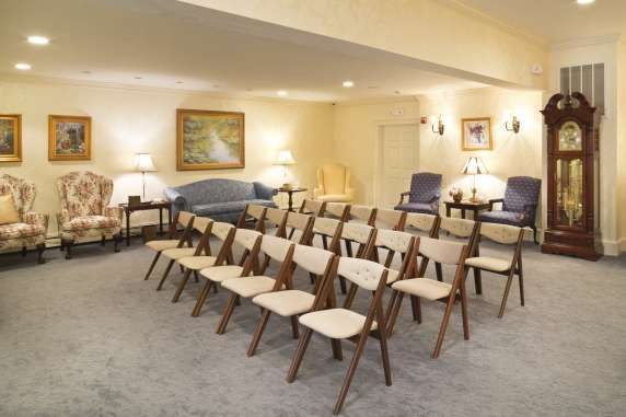 A funeral home viewing room with rows of chairs, sofas, paintings, and a grandfather clock.