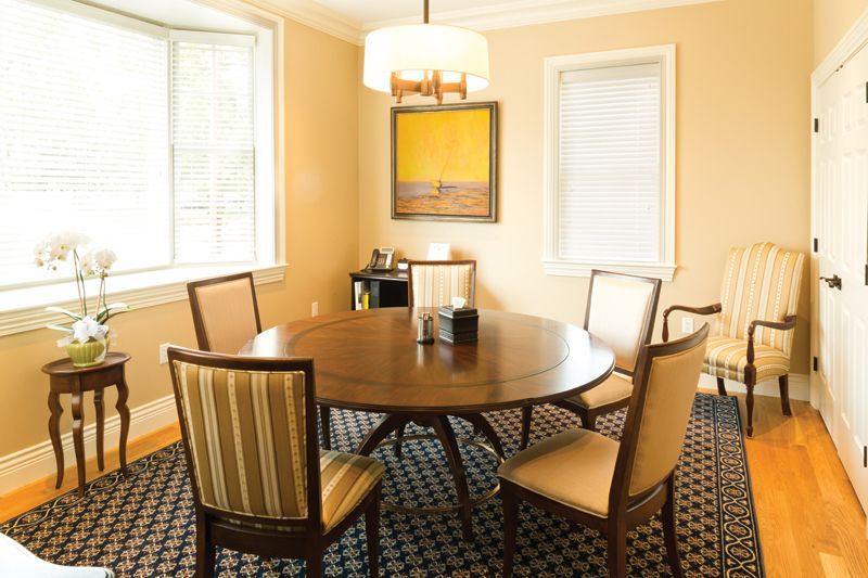 Dining room with round wooden table, chairs, artwork, and window with blinds.
