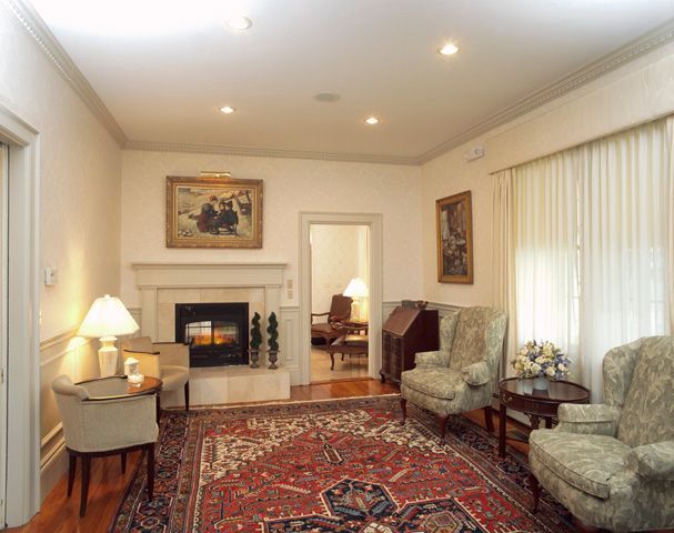 Cozy living room with fireplace, area rug, armchairs, paintings, and a doorway leading to another room.
