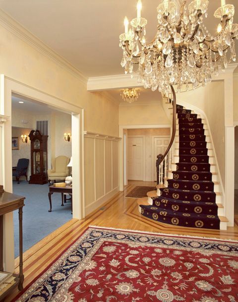 Elegant foyer with curved staircase, red carpet, and crystal chandelier.
