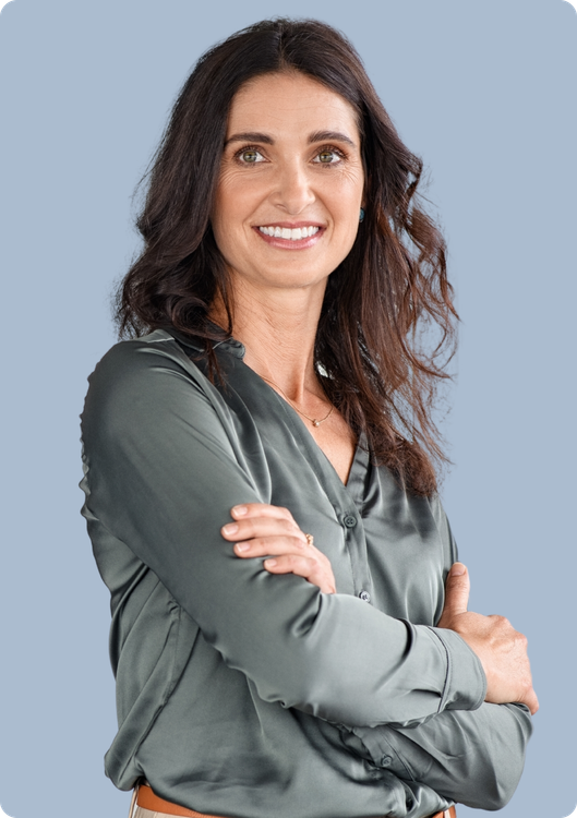 A person with long, dark hair wears a gray satin button-down shirt and smiles while standing with arms crossed.