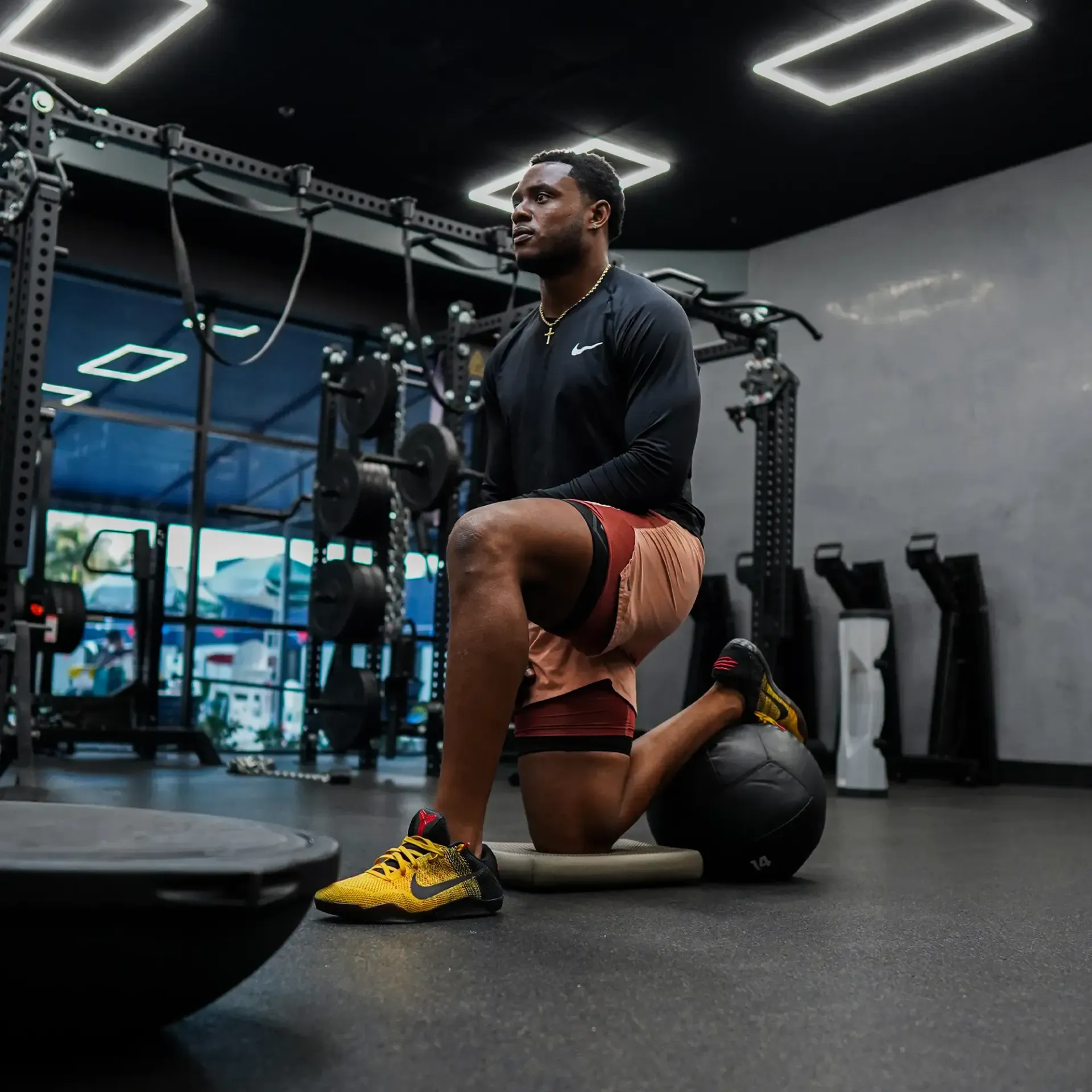 Man in a gym doing a lunge, one knee on a mat with a medicine ball.