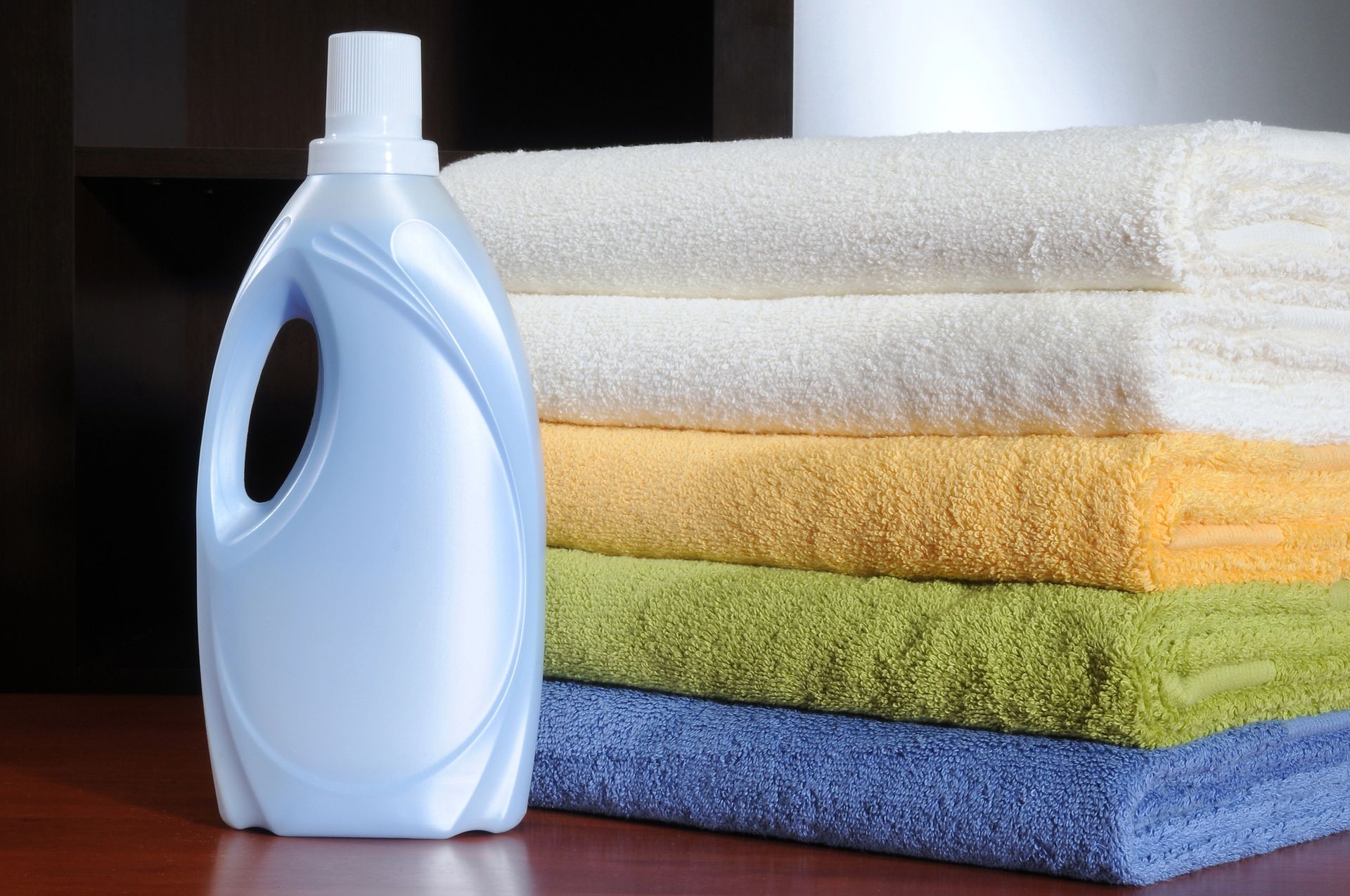 Botella de detergente para ropa junto a toallas blancas, beige, amarillas, verdes y azules cuidadosamente dobladas.