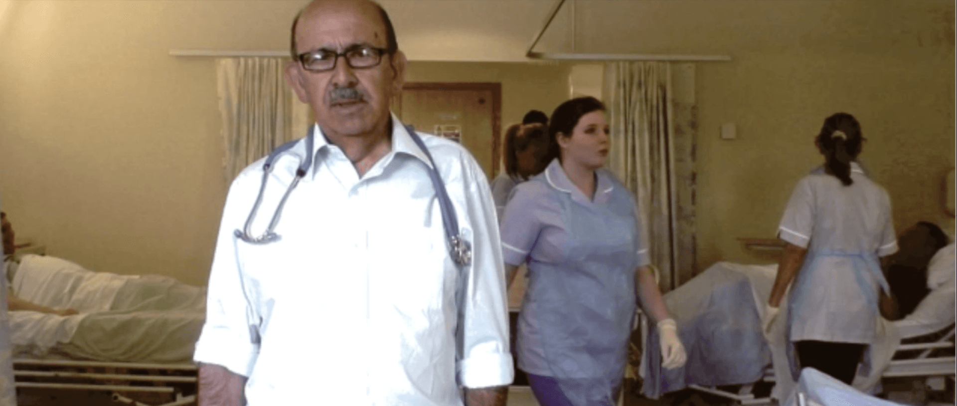 A hospital room. A doctor with a stethoscope around his neck is looking at the camera. There are 3 nurses and 2 patients in the background